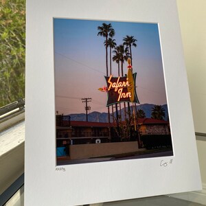 May include: A vintage neon sign for the Safari Inn motel, with palm trees in the background. The sign is lit up in pink and orange, with the words "Safari Inn" in a stylised font.