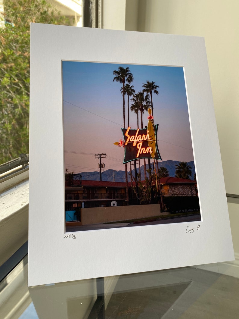 May include: A vintage neon sign for the Safari Inn motel, with palm trees in the background. The sign is lit up in pink and orange, with the words "Safari Inn" in a stylised font.