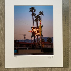 May include: A vintage neon sign for the "Safari Inn" motel, with palm trees in the background. The sign is lit up in orange and yellow, with a green and white surfboard design.