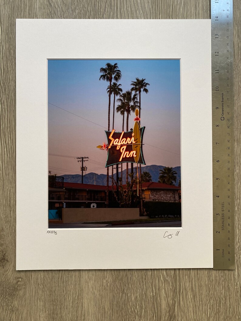May include: A vintage neon sign for the "Safari Inn" motel, with palm trees in the background. The sign is lit up in orange and yellow, with a green and white surfboard design.