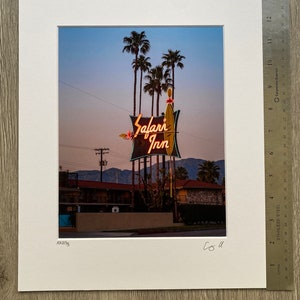 May include: A vintage neon sign for the Safari Inn motel, with palm trees in the background. The sign is lit up in orange and yellow, with the words "Safari Inn" in a stylised font.