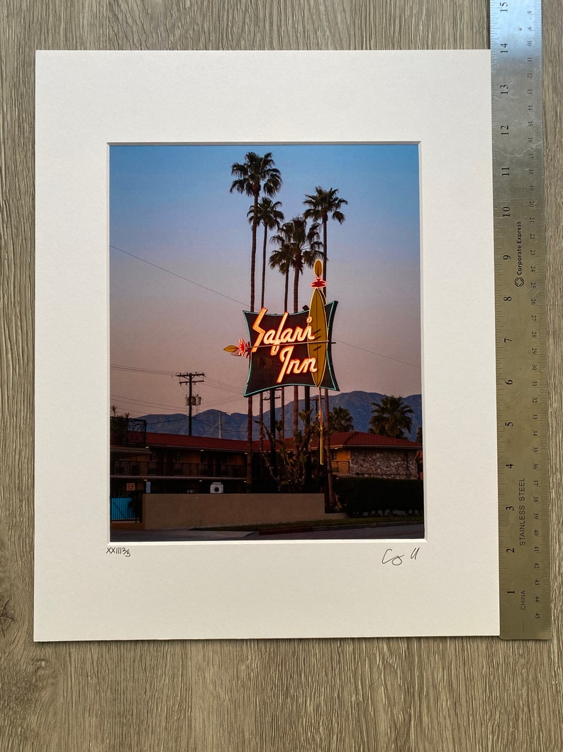 May include: A vintage neon sign for the Safari Inn motel, with palm trees in the background. The sign is lit up in orange and yellow, with the words "Safari Inn" in a stylised font.