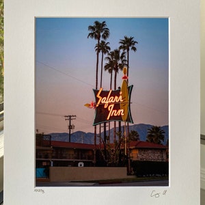 May include: A vintage motel sign with neon lights that reads "Safari Inn" against a twilight sky. Palm trees are in the foreground and a mountain range is in the background.