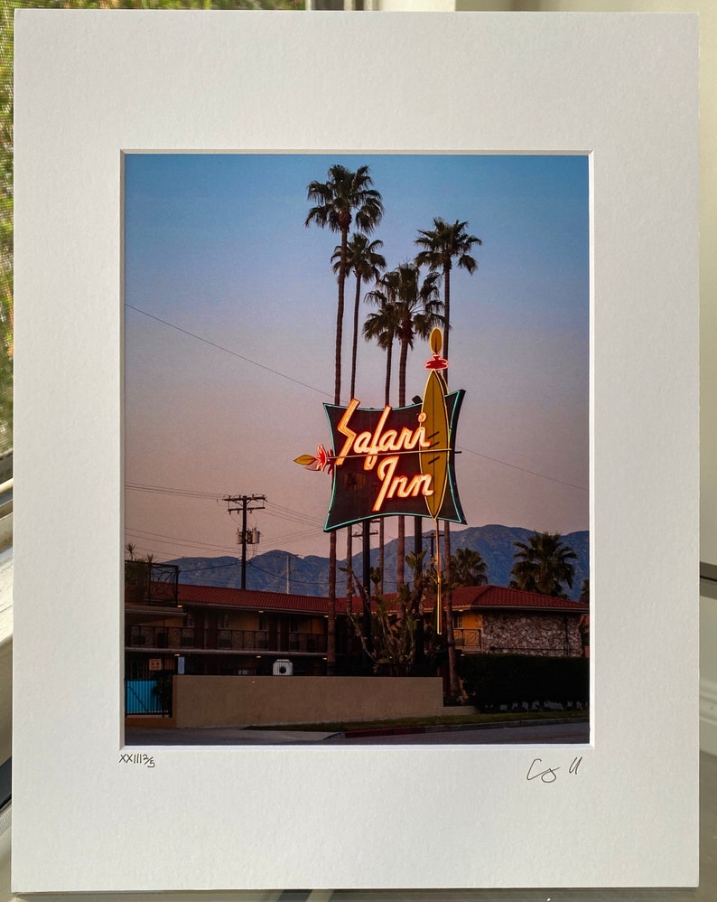 May include: A vintage motel sign with neon lights that reads "Safari Inn" against a twilight sky. Palm trees are in the foreground and a mountain range is in the background.