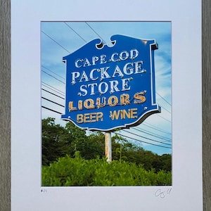 May include: A blue and gold sign with white lettering reads "Cape Cod Package Store Liquors Beer Wine". The sign is mounted on a rusty pole and surrounded by green foliage.