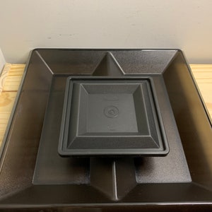 Puede incluir: Una bandeja cuadrada de plástico negro con una bandeja cuadrada negra más pequeña en su interior. La bandeja más pequeña tiene un borde elevado alrededor del borde.