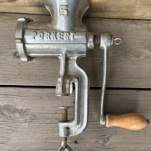 May include: A silver metal hand-crank meat grinder with a wooden handle. The grinder has a clamp to attach it to a surface. The grinder has the number "5" and the word "PORKERT" printed on it.
