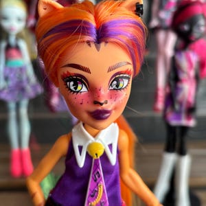 May include: A doll with orange and purple hair, cat ears, and a purple and white outfit. The doll has detailed facial features, including dark purple lipstick and freckles. Other dolls are visible in the background.