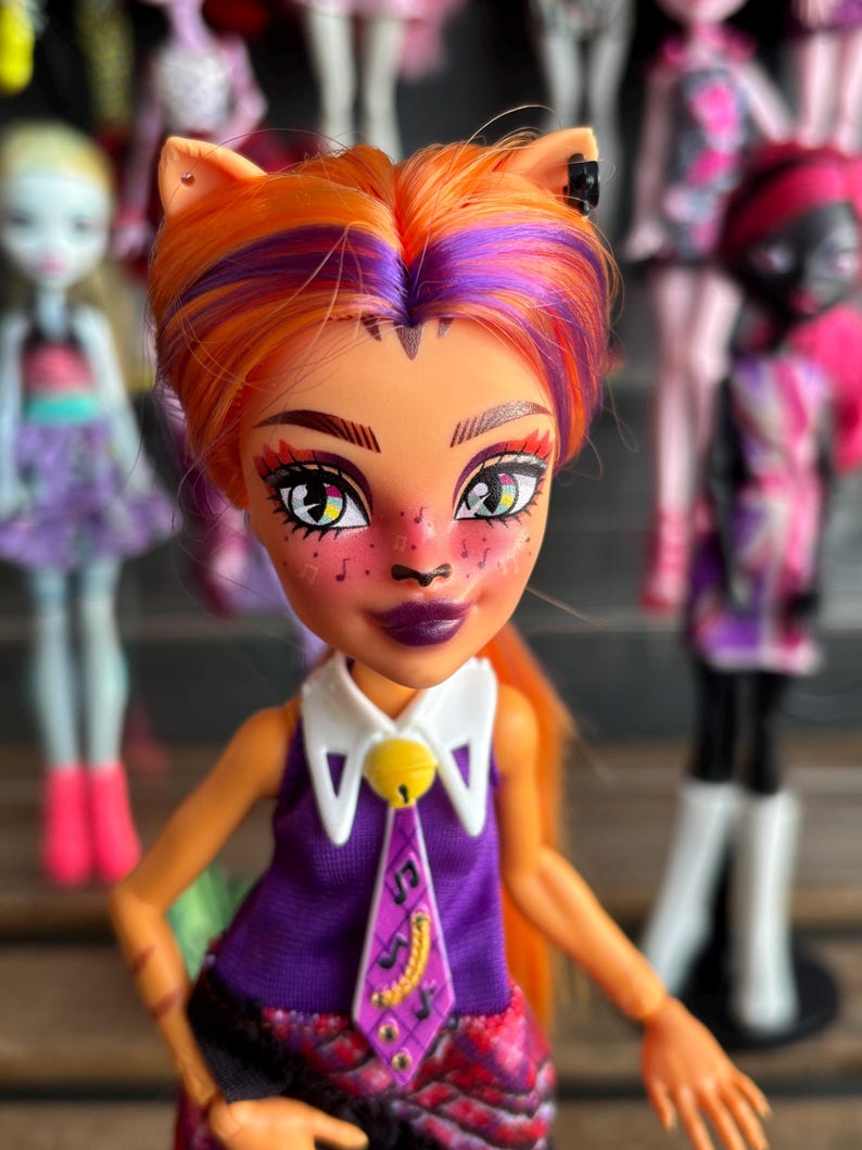 May include: A doll with orange and purple hair, cat ears, and a purple and white outfit. The doll has detailed facial features, including dark purple lipstick and freckles. Other dolls are visible in the background.