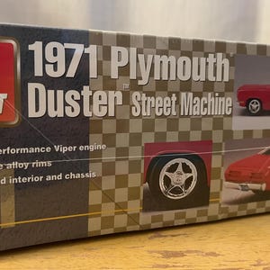 Op de afbeelding: Een modelbouwdoos voor een 1971 Plymouth Duster Street Machine. De doos toont een rode Duster-modelwagen, een close-up van de wielen van de auto en een gedetailleerde beschrijving van de kenmerken van de kit.