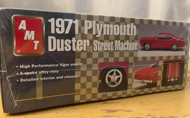 Op de afbeelding: Een modelbouwdoos voor een 1971 Plymouth Duster Street Machine. De doos toont een rode Duster-modelwagen, een close-up van de wielen van de auto en een gedetailleerde beschrijving van de kenmerken van de kit.