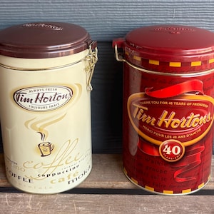 Puede incluir: Dos latas de café Tim Hortons. Una es de color crema con tapa marrón y detalles dorados, con el texto "ALWAYS FRESH" y "TOUJOURS FRAIS". La otra es roja con tapa a juego y el texto "THANK YOU FOR 40 YEARS OF FRIENDSHIP".