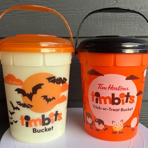 Puede incluir: Dos cubos de Halloween de Tim Hortons. Uno es blanquecino con tapa y asa naranja, con un gráfico de luna y murciélagos. El otro es naranja con tapa y asa negra, con un gráfico de Tim Hortons Timbits.