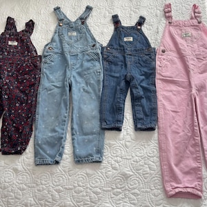 K&ouml;nnte beinhalten: Vier Kinder-Latzhosen in verschiedenen Farben und Mustern. Es gibt ein Paar mit dunklem Blumenmuster, zwei Jeans-Paare und ein rosa Cord-Paar. Alle Latzhosen haben verstellbare Tr&auml;ger und Taschen.