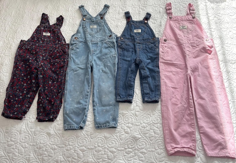 K&ouml;nnte beinhalten: Vier Kinder-Latzhosen in verschiedenen Farben und Mustern. Es gibt ein Paar mit dunklem Blumenmuster, zwei Jeans-Paare und ein rosa Cord-Paar. Alle Latzhosen haben verstellbare Tr&auml;ger und Taschen.