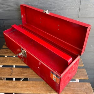 May include: Open red metal toolbox with a hinged lid and a tray. The toolbox has a silver latch and a small label with text and a maple leaf. The toolbox is sitting on a wooden surface.