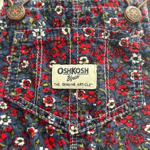 K&ouml;nnte beinhalten: Nahaufnahme einer blauen Cord-Latzhose mit einem Blumenmuster aus roten und wei&szlig;en Bl&uuml;ten. Die Latzhose hat eine Vordertasche mit dem Etikett "OSHKOSH B'gosh". Die Tr&auml;ger werden von bronzefarbenen Metallverschl&uuml;ssen gehalten.