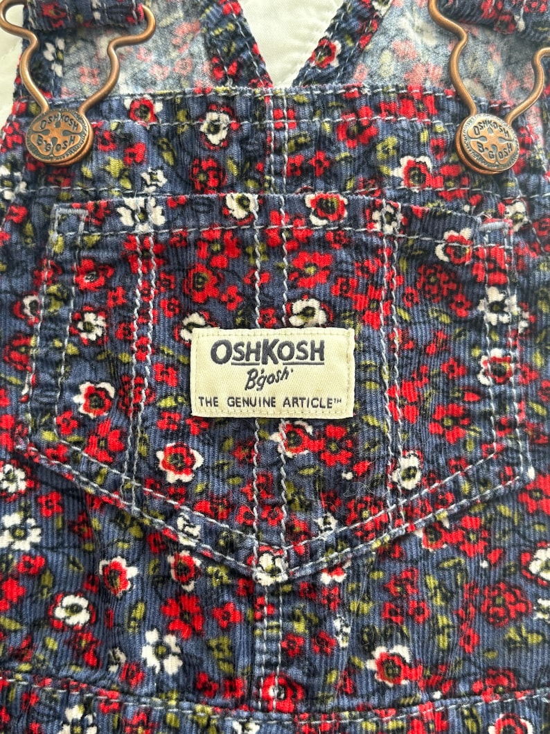 K&ouml;nnte beinhalten: Nahaufnahme einer blauen Cord-Latzhose mit einem Blumenmuster aus roten und wei&szlig;en Bl&uuml;ten. Die Latzhose hat eine Vordertasche mit dem Etikett "OSHKOSH B'gosh". Die Tr&auml;ger werden von bronzefarbenen Metallverschl&uuml;ssen gehalten.