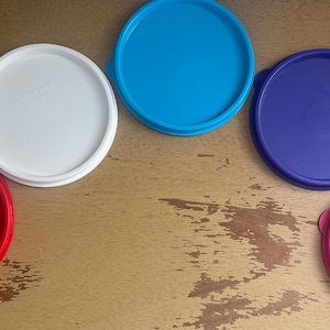 May include: Five plastic lids in different colors: red, white, blue, purple, and pink. The lids are arranged in a semi-circle on a wooden surface.