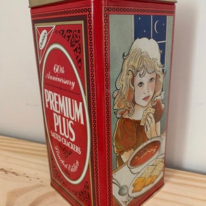 Puede incluir: Una lata rectangular de metal rojo para galletas saladas Christie's Premium Plus. El frente presenta el texto 60th Anniversary Premium Plus Salted Crackers Limited Edition 450g. El lado muestra una ilustración de un niño comiendo galletas y sopa.