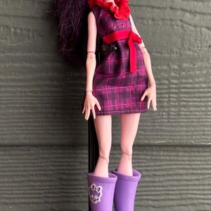 May include: A doll with dark purple hair, wearing a plaid dress with a red bow and purple boots. The doll has pale skin and red lipstick, standing on a black stand.