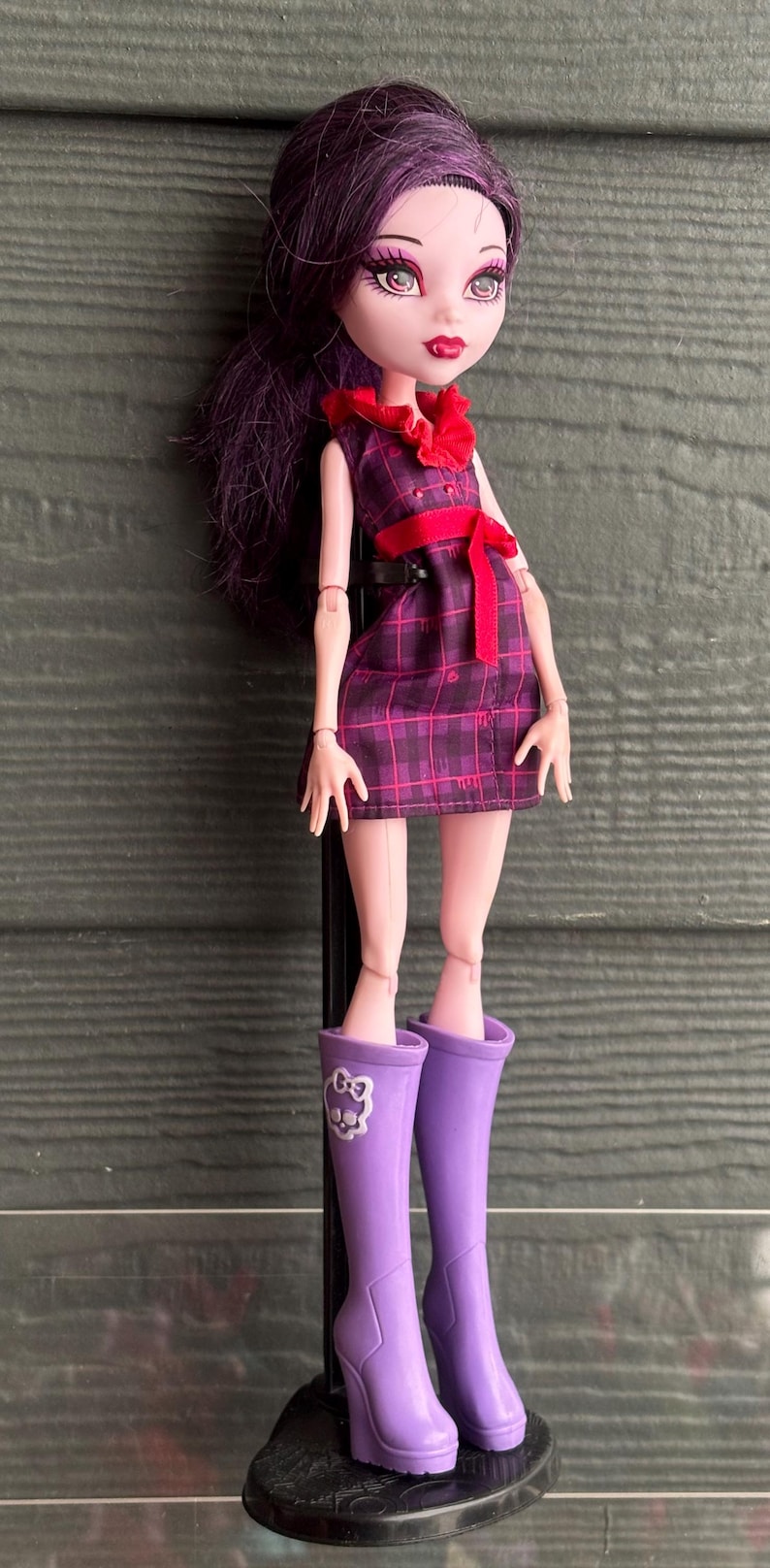 May include: A doll with dark purple hair, wearing a plaid dress with a red bow and purple boots. The doll has pale skin and red lipstick, standing on a black stand.