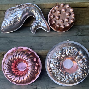 May include: Four copper cake pans, two round, one shaped like a fish, and one with a swirl design. The pans are all shiny and have a vintage look.