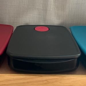 May include: Three rectangular food storage containers in red, black, and teal. Each container has a matching colored lid with a circular button. The containers are made of plastic and sit on a wooden surface.