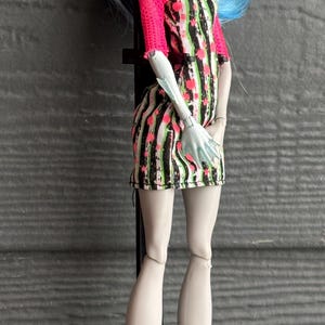 May include: A doll with blue and yellow hair, wearing a black, white, and pink striped dress with pink sleeves. The doll has roller skates with black boots and pink and green wheels. The doll is on a black stand.