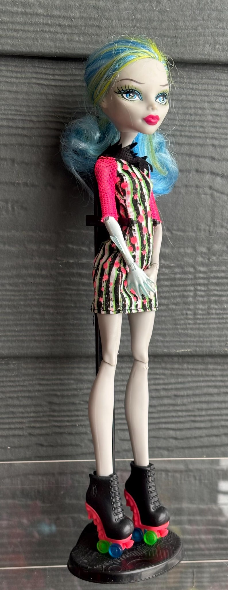 May include: A doll with blue and yellow hair, wearing a black, white, and pink striped dress with pink sleeves. The doll has roller skates with black boots and pink and green wheels. The doll is on a black stand.