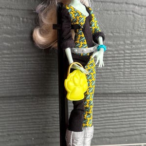 May include: A doll with pale green skin, cat-like ears, and light blonde hair. She wears a black and yellow patterned outfit, silver boots with yellow heels, and carries a yellow purse. The doll is on a black stand.