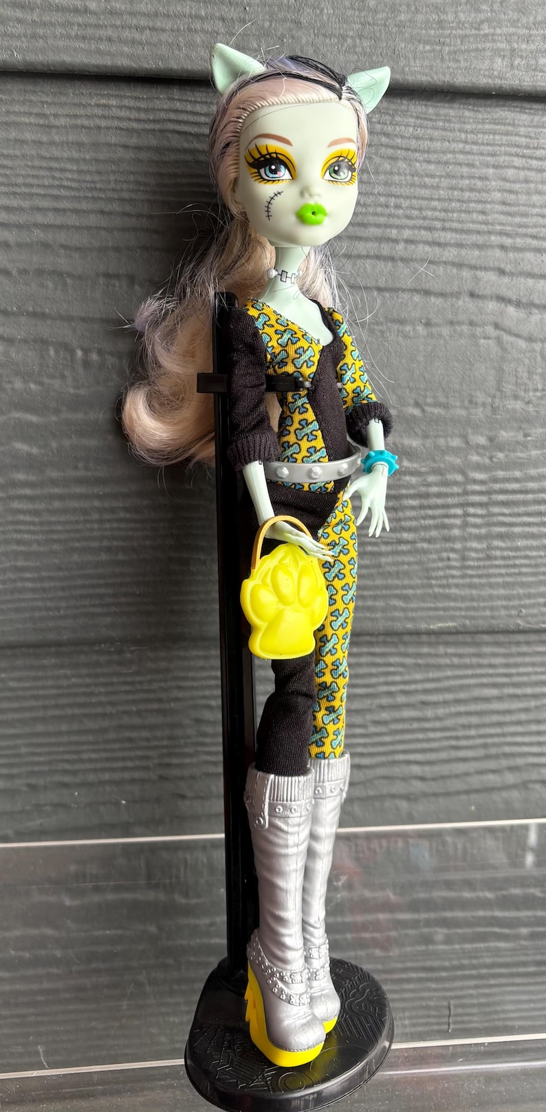 May include: A doll with pale green skin, cat-like ears, and light blonde hair. She wears a black and yellow patterned outfit, silver boots with yellow heels, and carries a yellow purse. The doll is on a black stand.