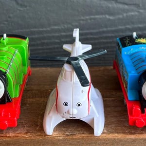 May include: A selection of toy trains and a helicopter. The trains are blue, green, and red, with black details. The helicopter is white with black rotor blades. All toys have smiling faces and are displayed on a wooden surface.