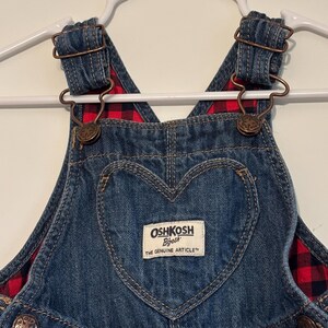 K&ouml;nnte beinhalten: Nahaufnahme einer blauen Jeans-Latzhose mit einer herzf&ouml;rmigen Tasche. Die Latzhose hat ein rot-schwarz kariertes Futter und Metallbeschl&auml;ge. Der Markenname "OshKosh B'gosh" ist auf einem rechteckigen Etikett zu sehen.