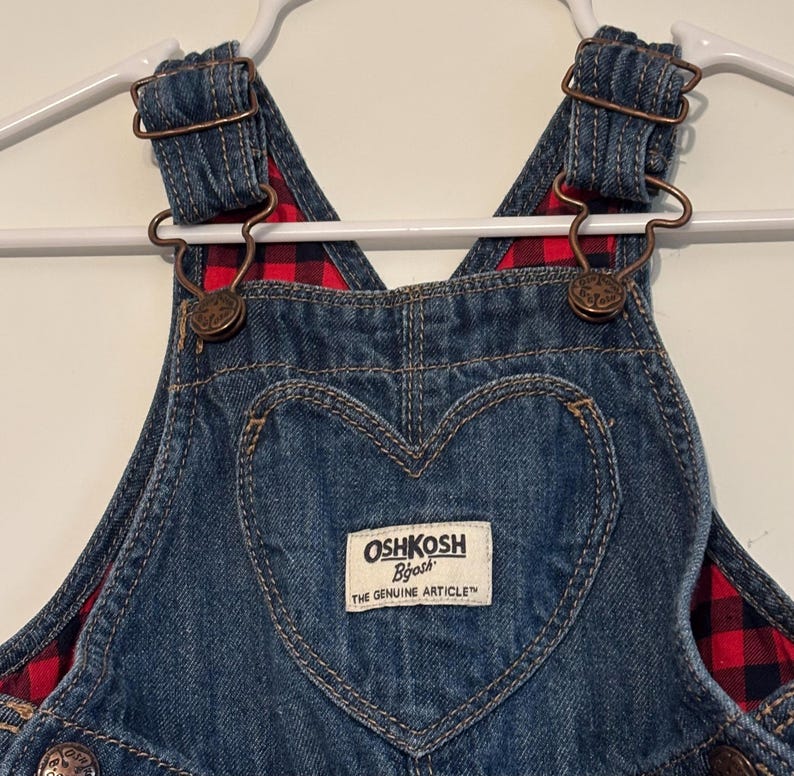 K&ouml;nnte beinhalten: Nahaufnahme einer blauen Jeans-Latzhose mit einer herzf&ouml;rmigen Tasche. Die Latzhose hat ein rot-schwarz kariertes Futter und Metallbeschl&auml;ge. Der Markenname "OshKosh B'gosh" ist auf einem rechteckigen Etikett zu sehen.