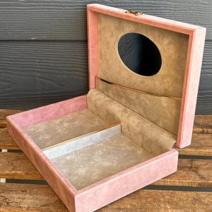 Könnte beinhalten: Geöffnete rosa Samt-Schmuckschatulle mit goldenem Verschluss und Zierleisten. Der Innenraum verfügt über einen Spiegel und Fächer, die mit einem hellbeigen Stoff ausgekleidet sind. Die Box steht auf einer Holzoberfläche.