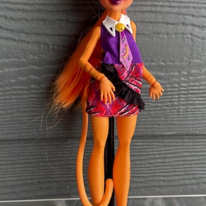 May include: A doll with orange skin, hair, and a tail. The doll has cat-like features, including ears and makeup. She wears a purple top, a plaid skirt, and pink boots. The doll is standing on a black stand.