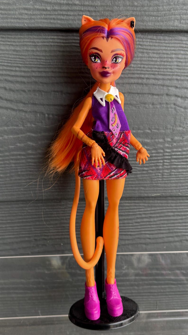 May include: A doll with orange skin, hair, and a tail. The doll has cat-like features, including ears and makeup. She wears a purple top, a plaid skirt, and pink boots. The doll is standing on a black stand.