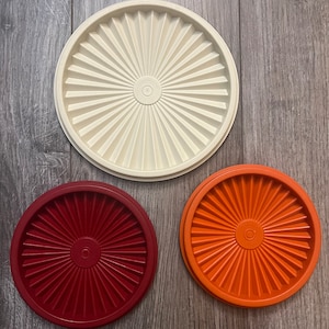 Puede incluir: Tres tapas de plástico para recipientes de almacenamiento en blanco, rojo y naranja. Cada tapa tiene un diseño circular con líneas radiales.