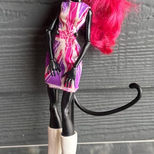 May include: A doll with black skin, cat ears, and bright pink hair. She wears a short dress with a pink and purple pattern and white boots with heels. A black tail extends from her back.