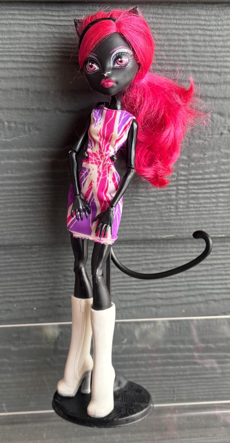May include: A doll with black skin, cat ears, and bright pink hair. She wears a short dress with a pink and purple pattern and white boots with heels. A black tail extends from her back.