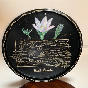 May include: A black metal tray with a gold outline and a map of South Dakota. The tray features a pasqueflower, the state flower, in the top right corner. The map shows the major cities and towns in the state, including Sioux Falls, Rapid City, and Aberdeen.