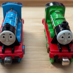 Op de afbeelding: Twee speelgoedtreinmotoren, één blauw en één groen, staan naast elkaar. De blauwe motor heeft een rode basis en de groene motor heeft een rode basis. Beide hebben lachende gezichten en zwarte schoorstenen. De treinen staan op een lichtbruin oppervlak.