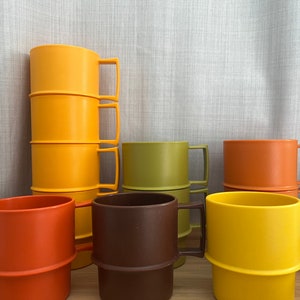 Könnte beinhalten: Eine Sammlung farbenfroher, stapelbarer Becher in Gelb-, Orange-, Grün- und Brauntönen. Die Becher haben einen einfachen, rechteckigen Griff und ein klassisches Design. Die Becher bestehen aus einem haltbaren Material.