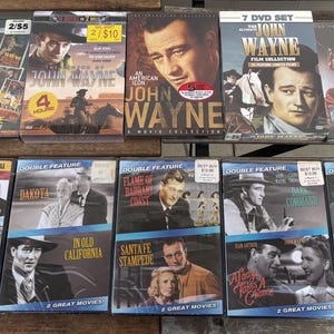 May include: A collection of John Wayne movie DVDs, featuring various titles like "The Sons of Katie Elder" and "Dakota." The covers showcase images of the actor and film titles, with some sets labeled as double features. The DVDs are displayed on a wooden surface.
