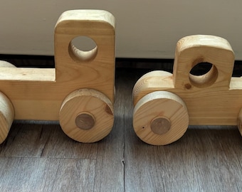 May include: Two wooden toy trucks. One truck has a flatbed, the other has a cab. Both are made of light-colored wood with rounded wheels and circular cutouts in the cab. The toys are on a wooden surface.