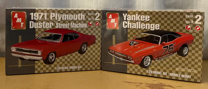 Op de afbeelding: Twee modelbouwsets voor auto's, een is een rode 1971 Plymouth Duster Street Machine en de andere is een rode Dodge Challenger met de tekst "Yankee Challenge" op de doos. Beide dozen zijn gemarkeerd met "AMT" en hebben een moeilijkheidsgraad van 10+.