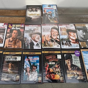 May include: A collection of John Wayne western movie VHS tapes, featuring titles like "The Shootist," "Rio Lobo," and "How the West Was Won." The tapes are displayed on a wooden surface, showcasing the classic film artwork.