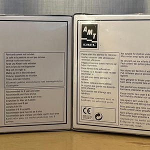 Op de afbeelding: Twee witte dozen met zwarte tekst en een zwart-wit logo met de tekst "AMT E-Z-BUILD". De dozen bevatten instructies voor het bouwen van een model. De dozen hebben waarschuwingen en veiligheidsinformatie erop gedrukt.