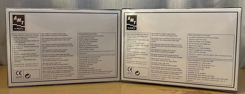 Op de afbeelding: Twee witte dozen met zwarte tekst en een zwart-wit logo met de tekst "AMT E-Z-BUILD". De dozen bevatten instructies voor het bouwen van een model. De dozen hebben waarschuwingen en veiligheidsinformatie erop gedrukt.